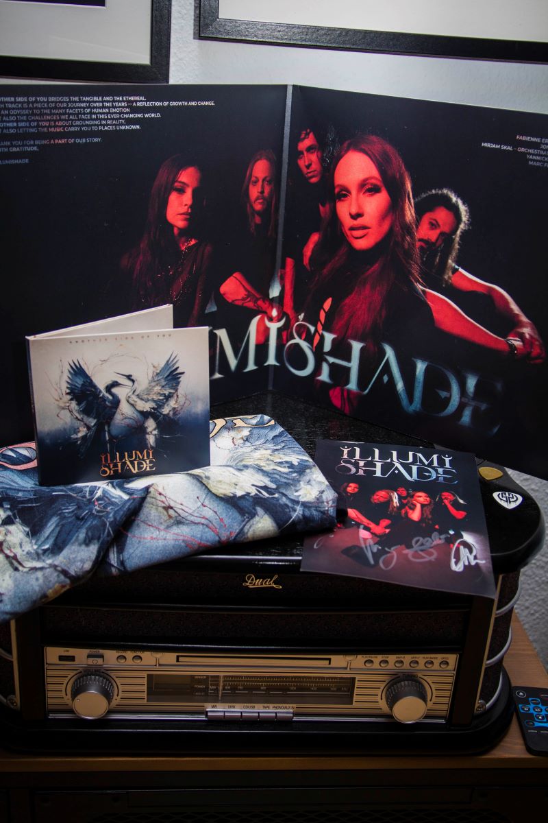 Photo shows the inside of the vinyl of ILLUMISHADE's album Another Side Of You in the background. It's placed on a record player. In front of it, there's the CD with a folded t-shirt underneath and next to the t-shirt there's a signature card