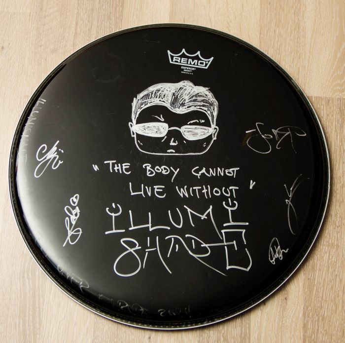 photo shows a black drumhead painted with silver edding. At the top there's the head of Neo from Matrix (stylised like a FunkoPop figure). Underneath there's the words "The body cannot live without ILLUMISHADE". Its surrounded by the signatures of the band members and at the edge there's (barely visible) a text saying that it's from the "Dark Waters over Europe" tour